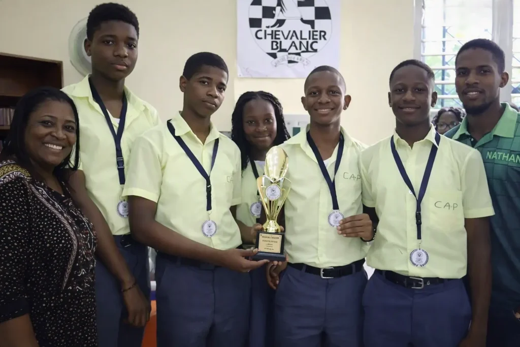 Élèves du CAP vainqueurs du tournoi de jeu d’échecs à Jacmel, portant des médailles et un trophée, accompagnés de leurs encadrants devant le panneau Chevalier Blanc.